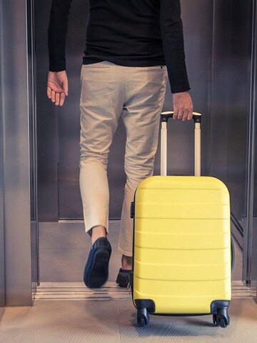 Person with a yellow suitcase entering an elevator, seen from behind, next to a stone wall.
