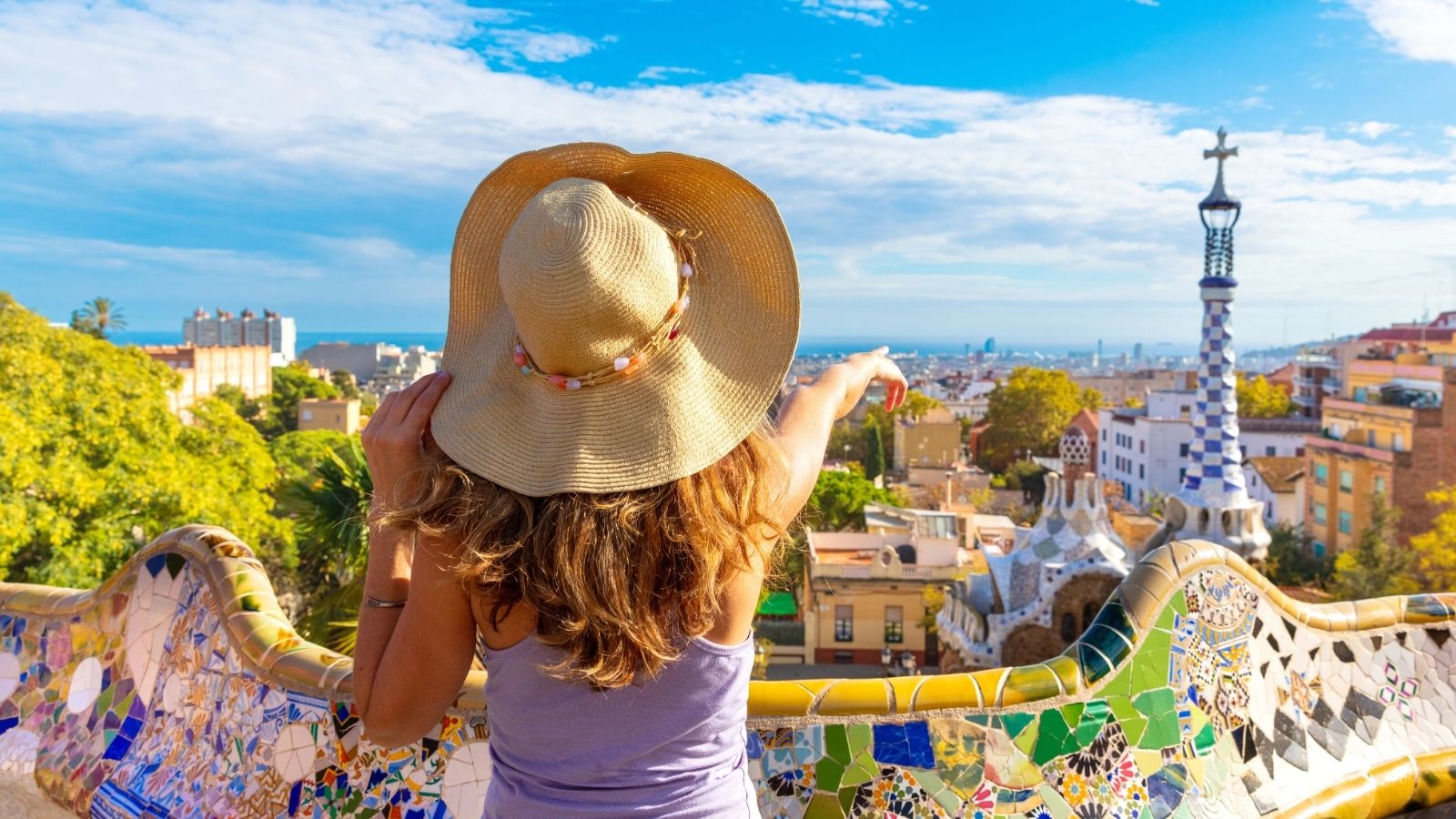 Woman in a sunhat points at a cityscape from a colorful mosaic terrace in a sunny park.