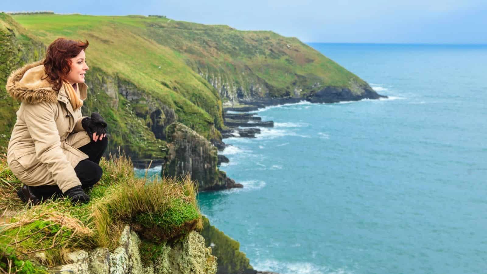 Woman in a tan coat sits on a grassy cliff overlooking the ocean and rocky coastline.
