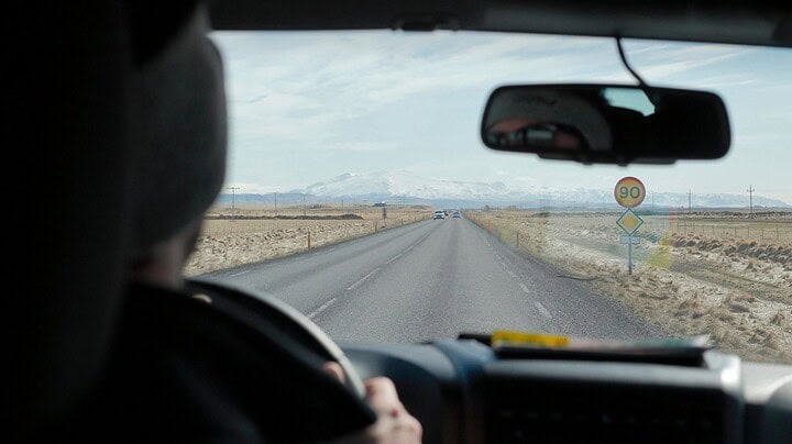 View from inside a car driving down a rural road toward distant mountains, speed limit sign shows 90 km/h.