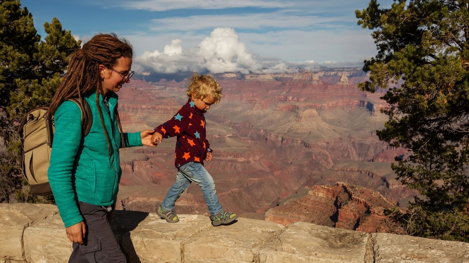 Adult and child holding hands, walking along a stone wall at the Grand Canyon with scenic views in the background.