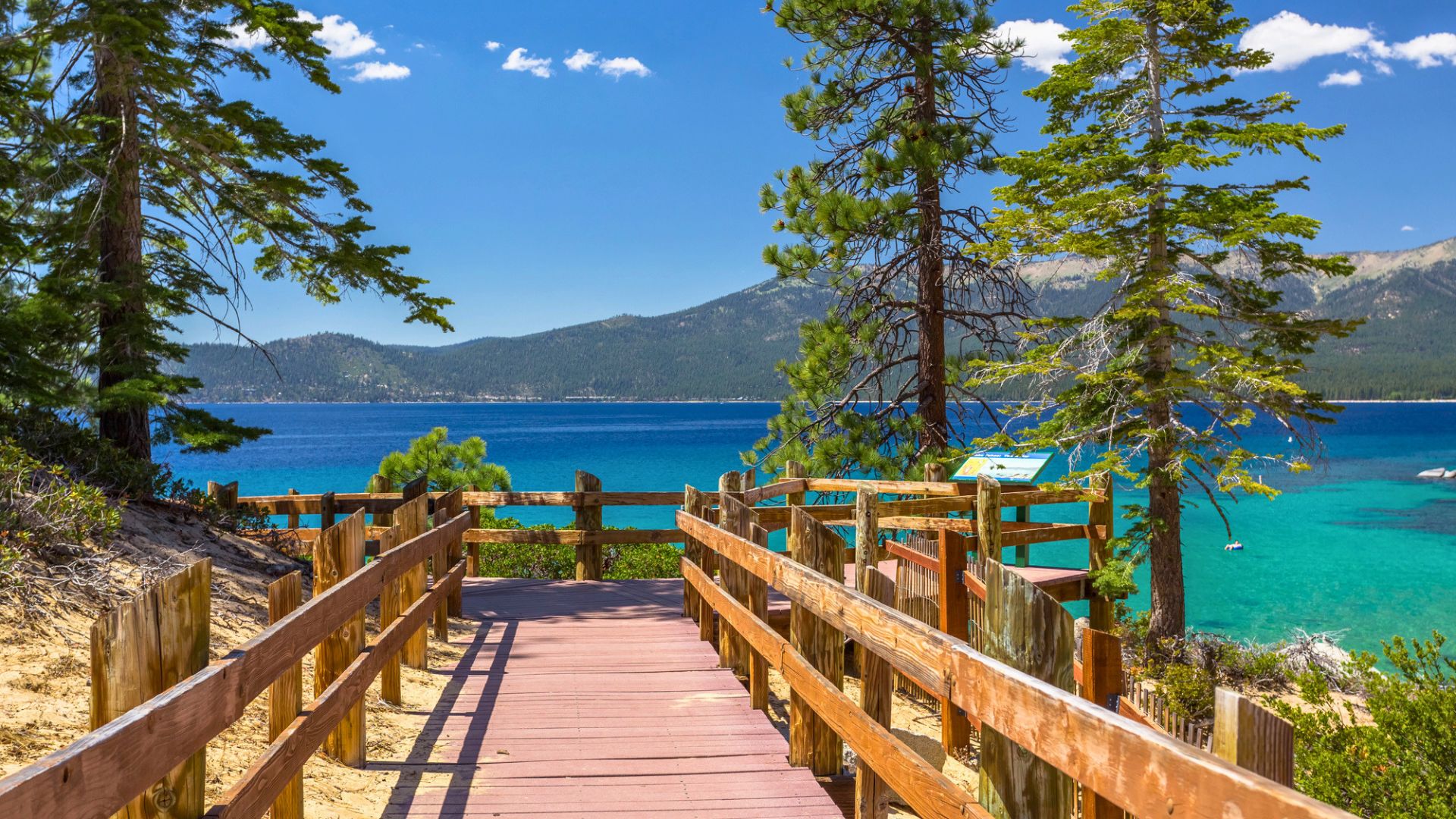 A wooden pathway leads to a clear blue lake, surrounded by trees and mountains under a sunny sky—the perfect setting for a family vacation to Lake Tahoe.
