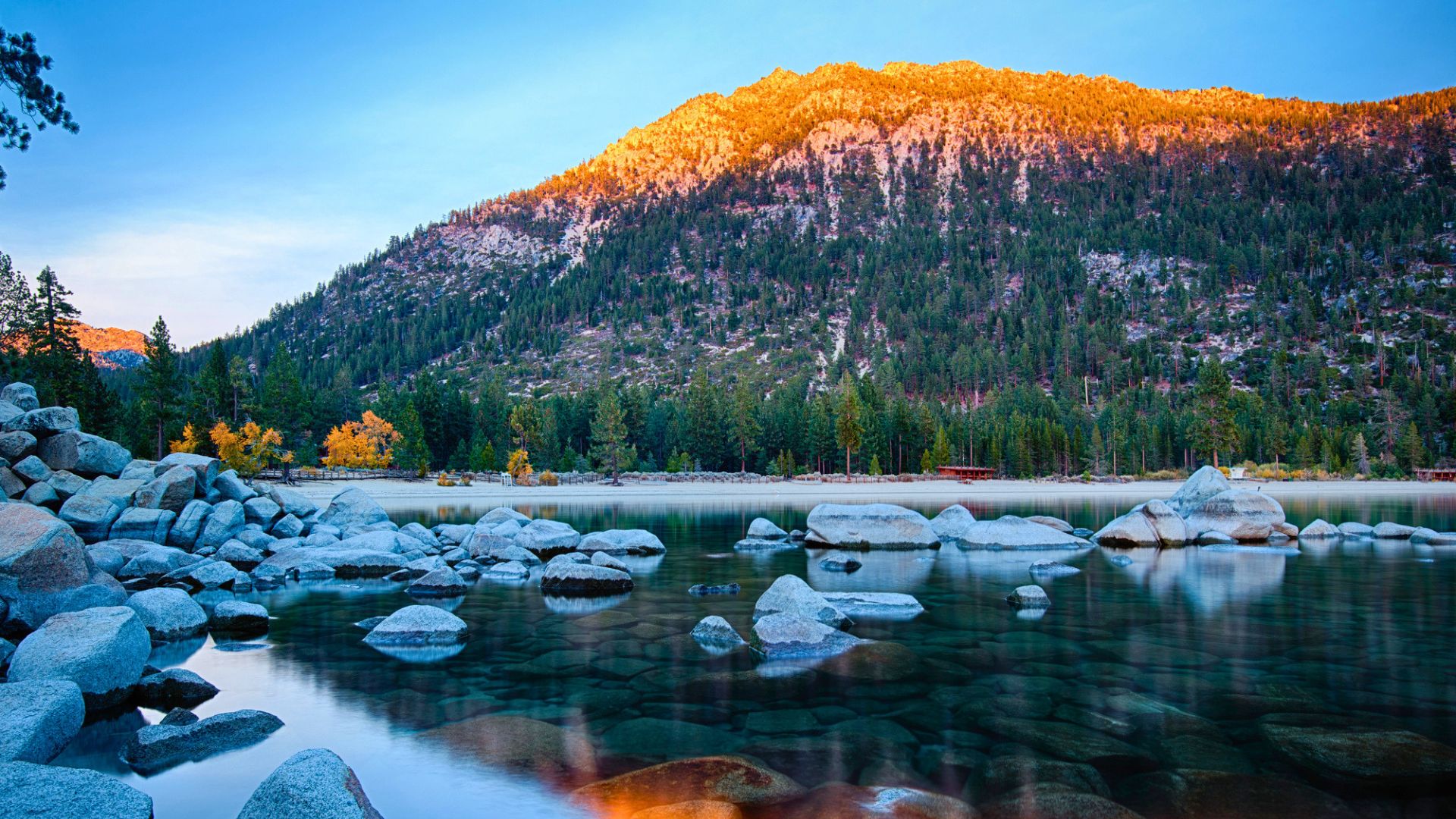 Clear lake with rocks in foreground, forest and mountain in background, mountain top lit by sunrise or sunset—a perfect scene for a family vacation to Lake Tahoe.