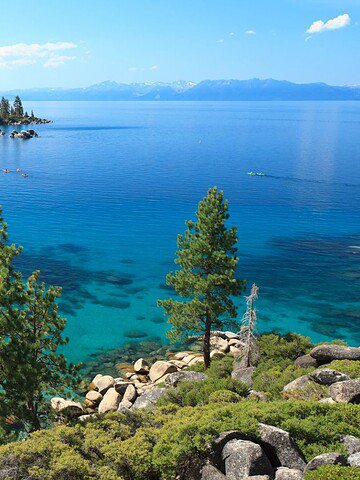 Clear blue lake surrounded by pine trees, rocky shorelines, and distant mountains under a sunny sky—perfect for a family vacation to Lake Tahoe.