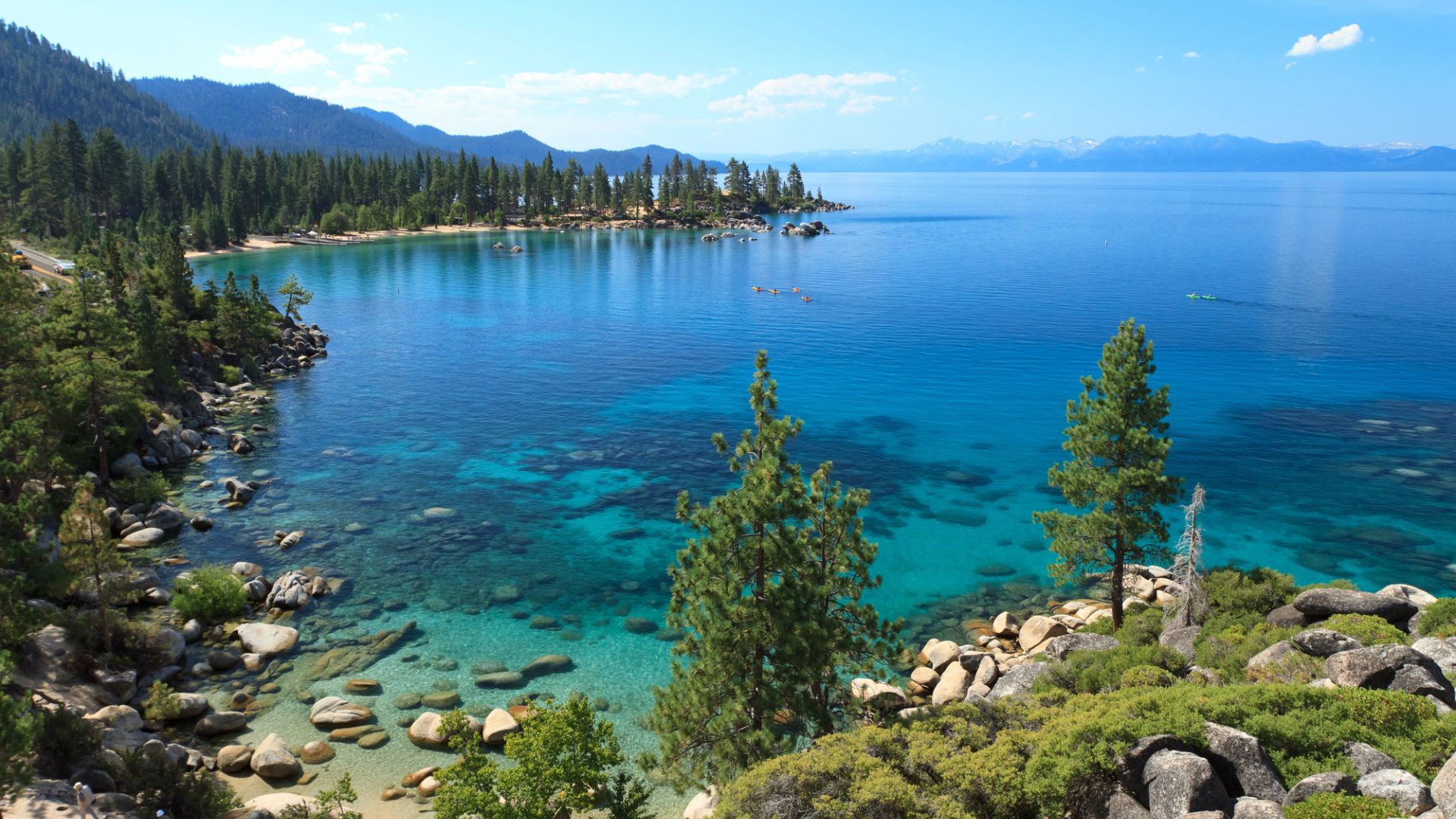 Clear blue lake surrounded by pine trees, rocky shorelines, and distant mountains under a sunny sky—perfect for a family vacation to Lake Tahoe.