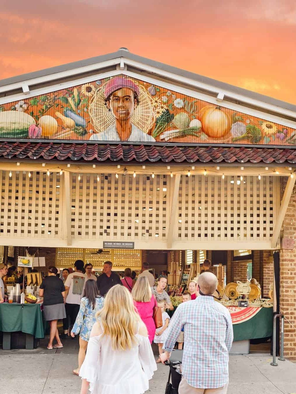 People walk toward a lively outdoor market at sunset, with a mural of a farmer and produce above the entrance—a perfect spot to explore if you’re looking for things to do in Charleston with kids.