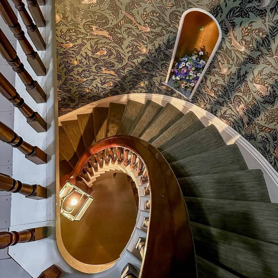 Curved staircase with green carpet, wooden railing, floral wallpaper, and a small alcove with flowers—perfect for those seeking the Best Places to Stay in Finger Lakes Near Wineries.