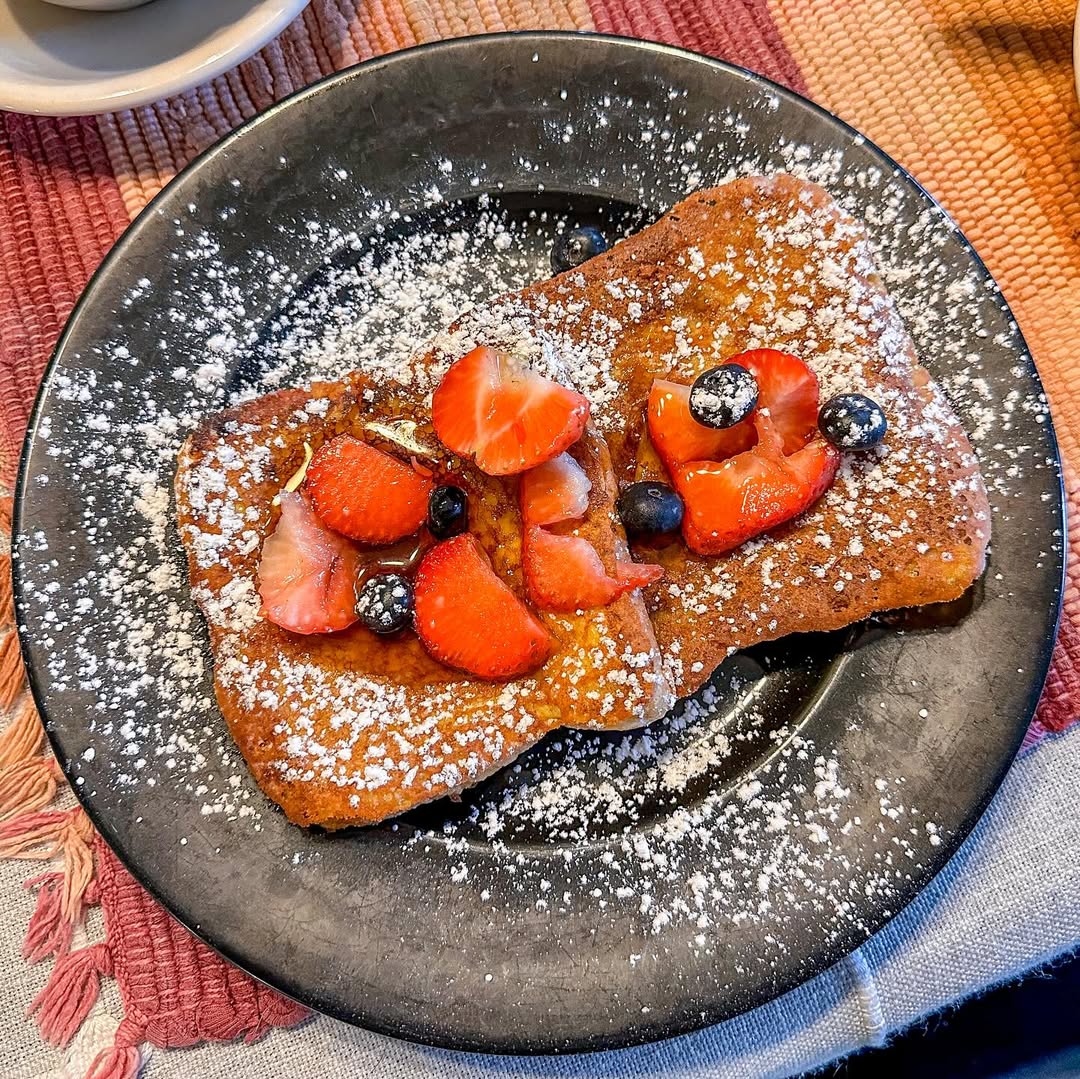 Two slices of French toast topped with strawberries, blueberries, and powdered sugar on a black plate—perfect for a morning at one of the best places to stay in Finger Lakes near wineries.