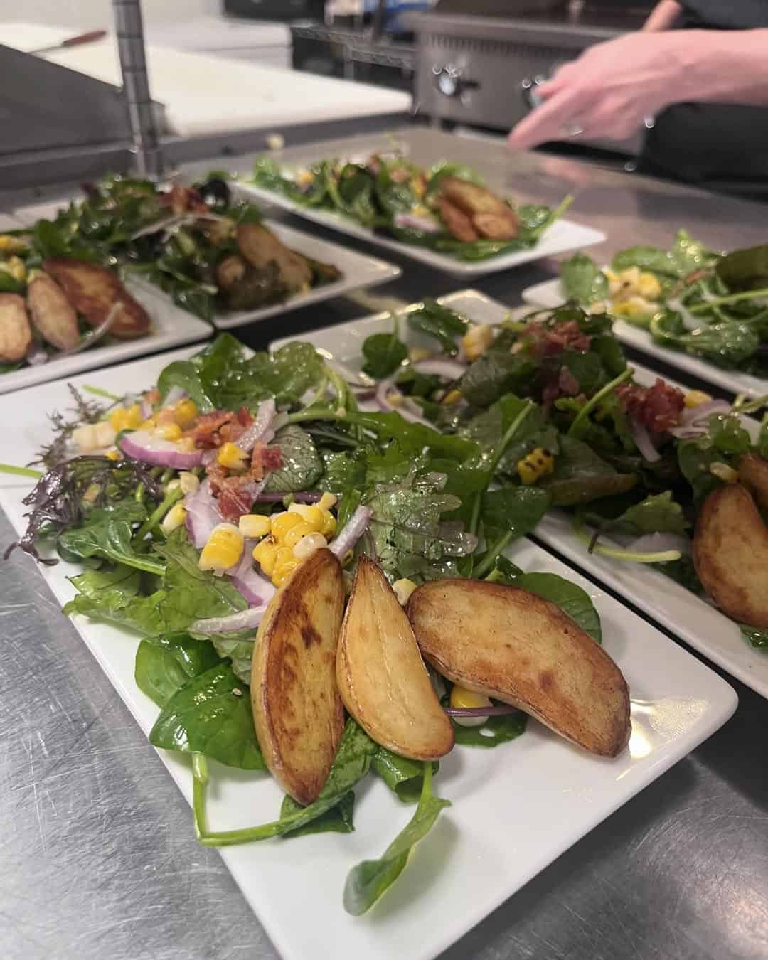 Plated salad with greens, corn, red onion, and potato wedges on a kitchen counter&mdash;just the kind of fresh dish you&rsquo;d find at Finger Lakes wineries with restaurants. Hand in the background adds a touch of lively ambiance.