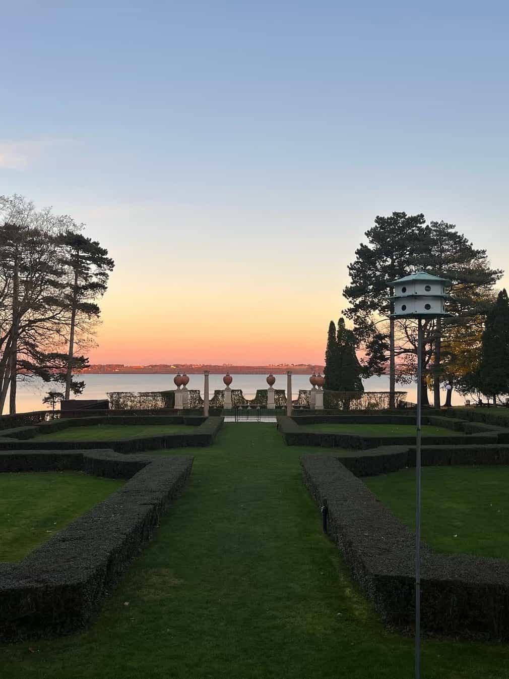 Manicured garden with trimmed hedges overlooking a lake at sunset, trees on both sides, and a birdhouse in view—capturing the serene charm of the Best Places to Stay in Finger Lakes Near Wineries.