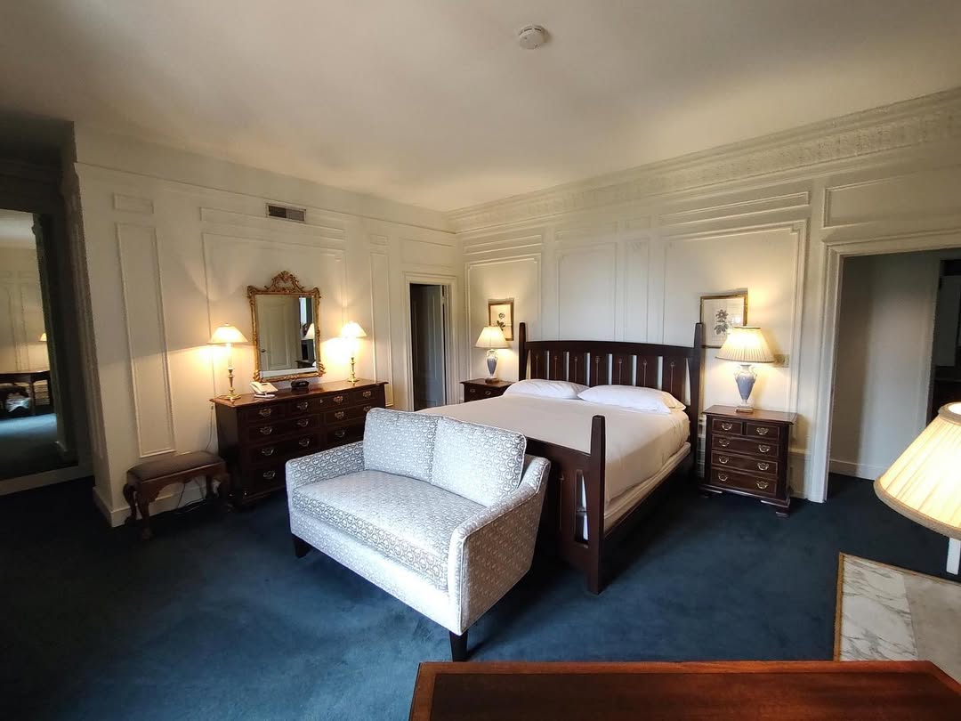 Elegant hotel room with a large bed, sofa, dressers, lamps, and mirrors on blue carpet flooring—one of the best places to stay in Finger Lakes near wineries.