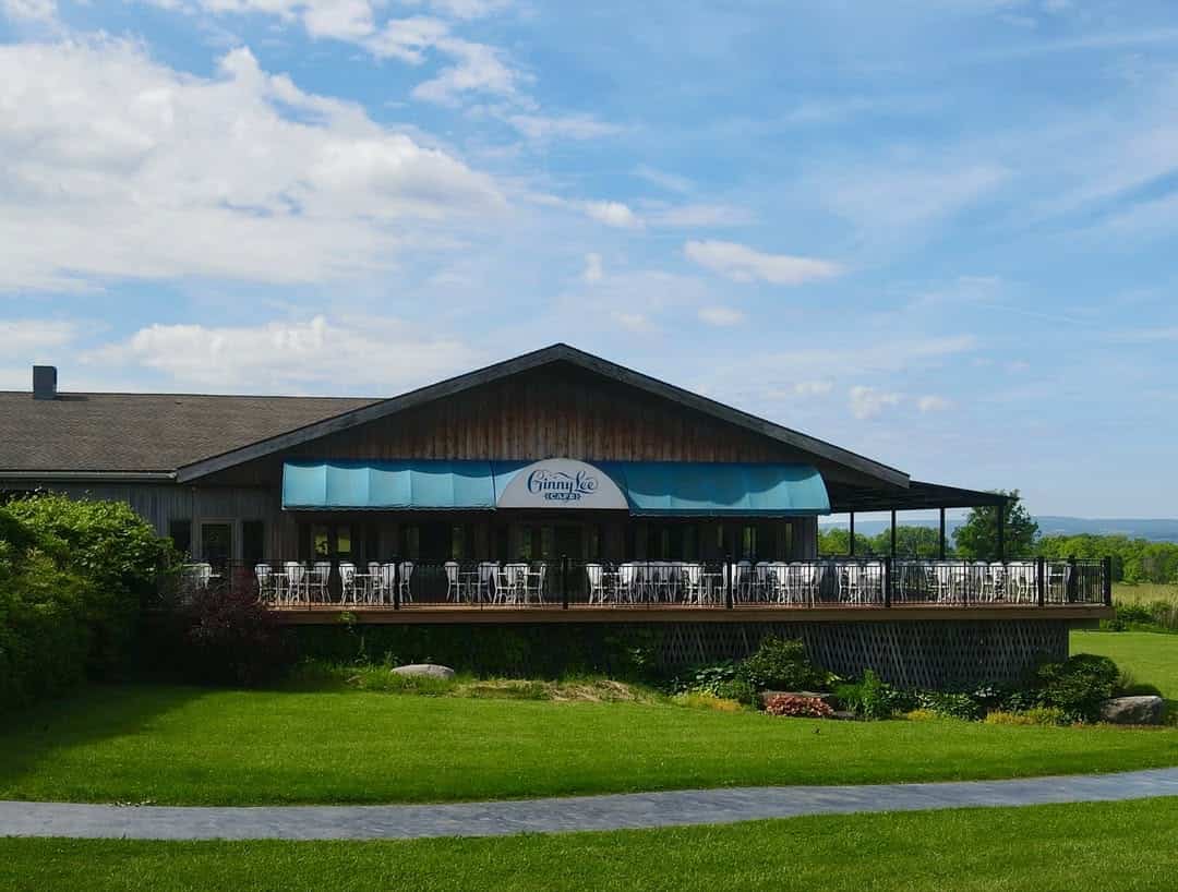 A rustic building with a blue awning and outdoor seating overlooks a green lawn under a partly cloudy sky, evoking the charm of Finger Lakes wineries with restaurants.