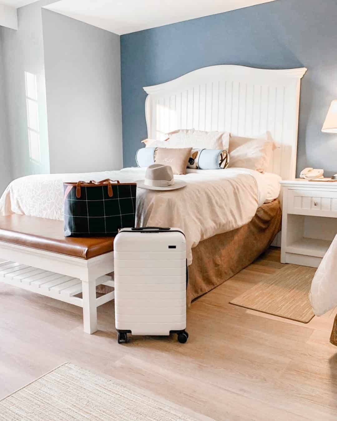 A suitcase, hat, and bag are placed next to a neatly made bed in a bright, cozy bedroom—perfect for those seeking the Best Places to Stay in Finger Lakes Near Wineries.