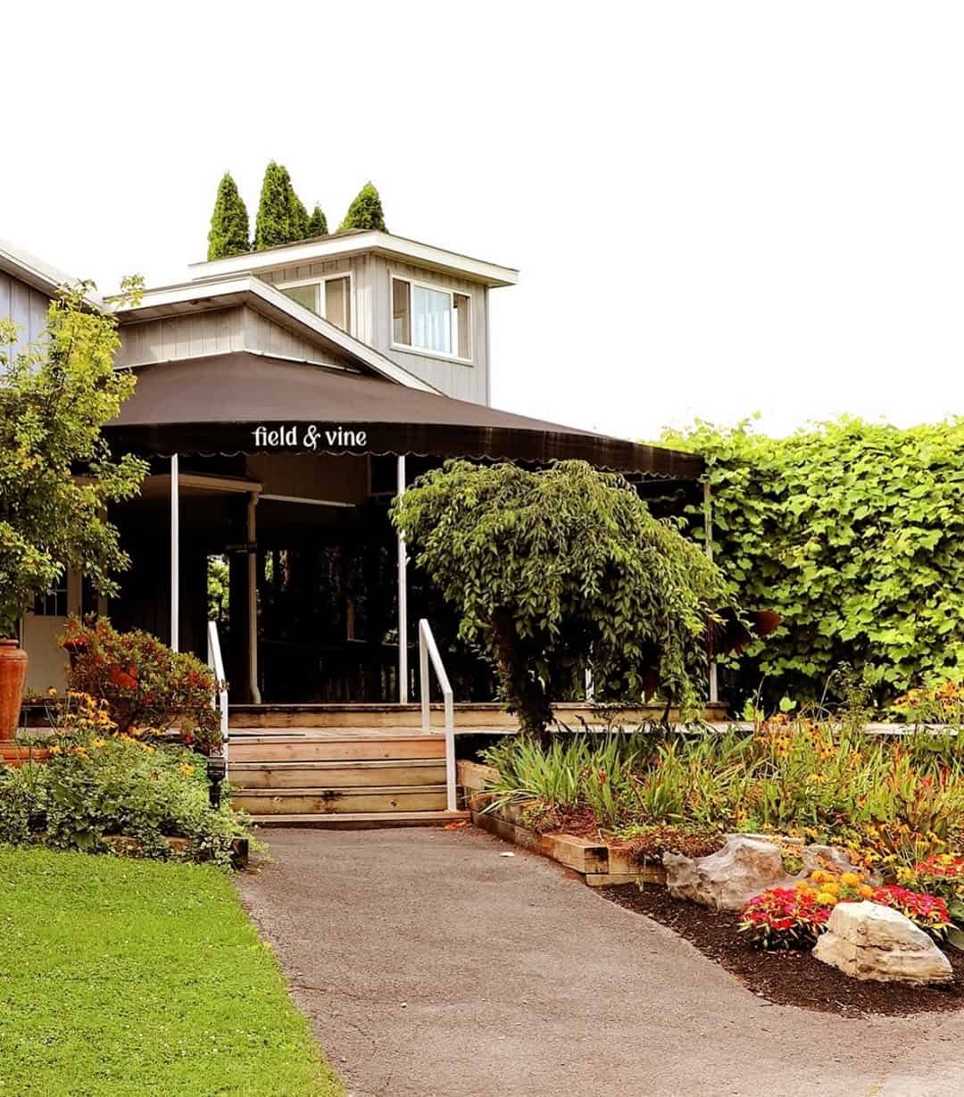 A garden path leads to the Field & Vine restaurant entrance with lush greenery and colorful flowers, reminiscent of the inviting settings found at Finger Lakes wineries with restaurants.