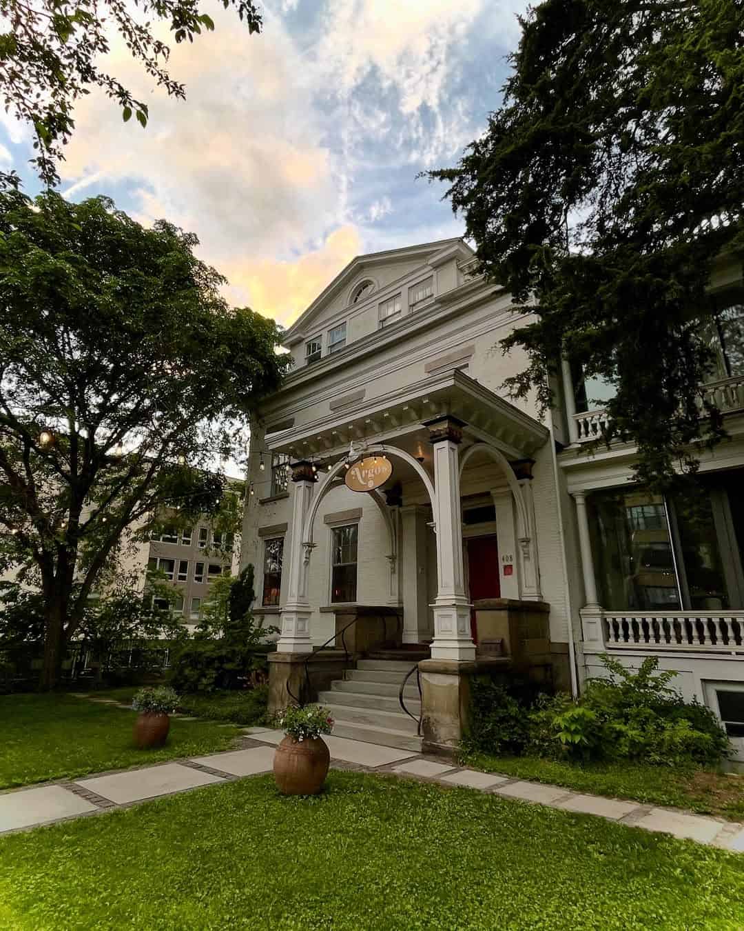 A grand white house with columns and a porch, surrounded by greenery and trees at sunset—one of the best places to stay in Finger Lakes near wineries, offering an inviting retreat amid scenic beauty.