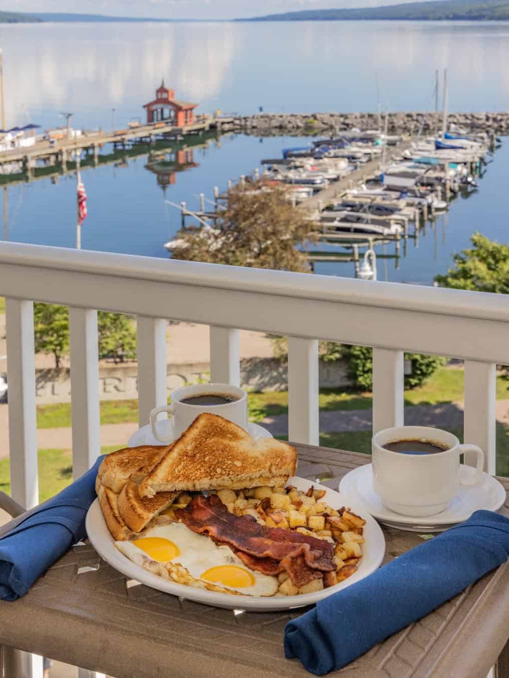 Enjoy a plate of eggs, bacon, toast, and potatoes with coffee on a balcony overlooking a marina and calm lake—one of the best places to stay in Finger Lakes near wineries for a relaxing breakfast experience.