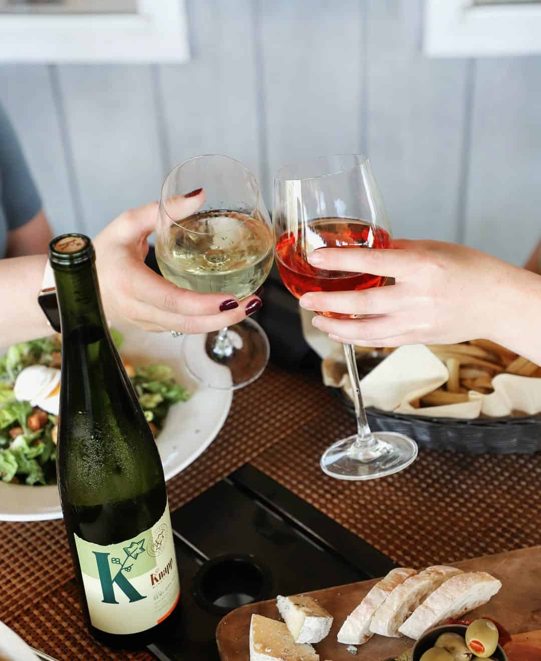 Two people clink glasses of white and ros&eacute; wine over a table with food, bread, and a wine bottle&mdash;capturing the spirit of Finger Lakes wineries with restaurants.