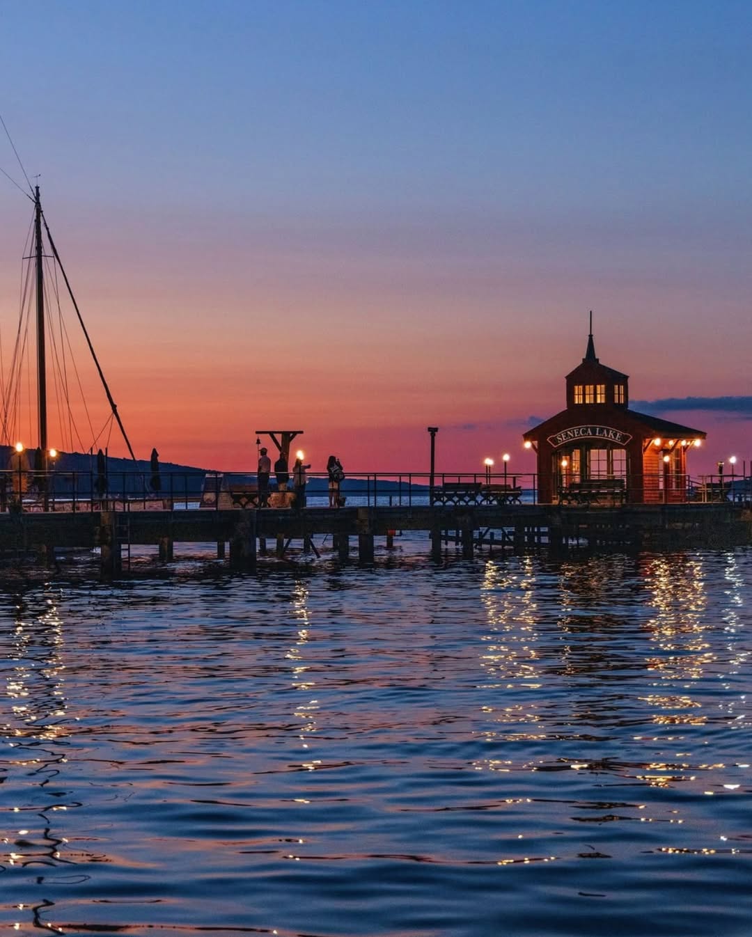 People standing on a pier at sunset, lights reflecting on calm water near a small building and sailboat—one of the best places to stay in Finger Lakes near wineries, offering serene views and unforgettable moments.