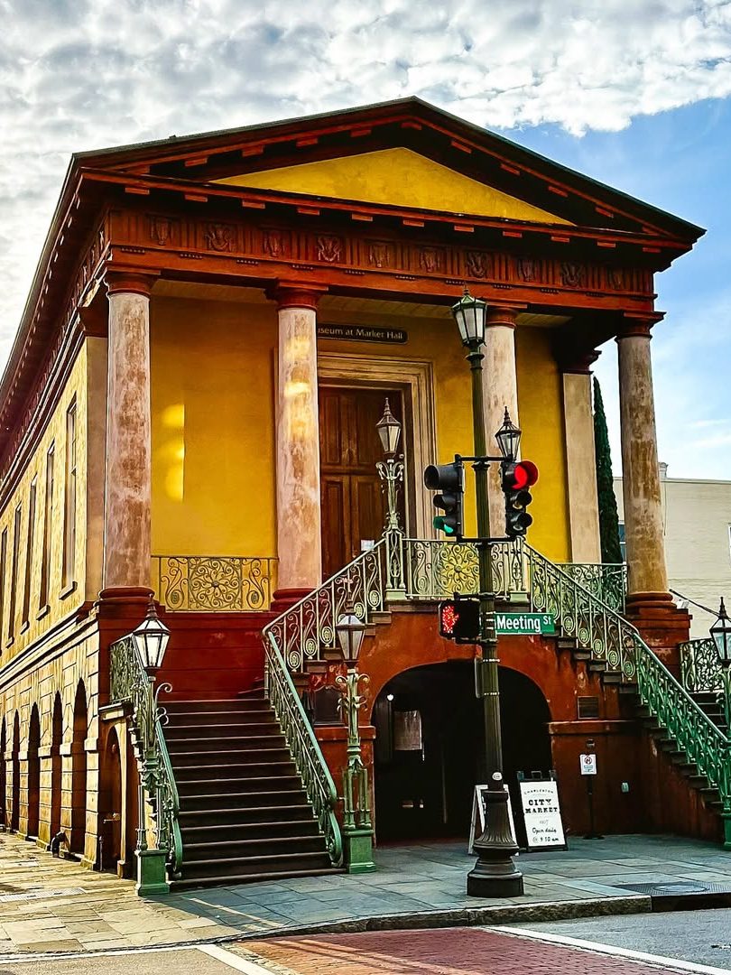 Historic two-story building with columns, twin staircases, and Meeting Street signs under a partly cloudy sky—a must-see for families exploring things to do in Charleston with kids.