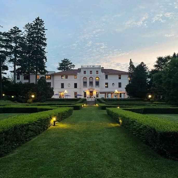 A grand mansion with trimmed hedges and a green lawn, lit by small garden lights at dusk—one of the best places to stay in Finger Lakes near wineries.