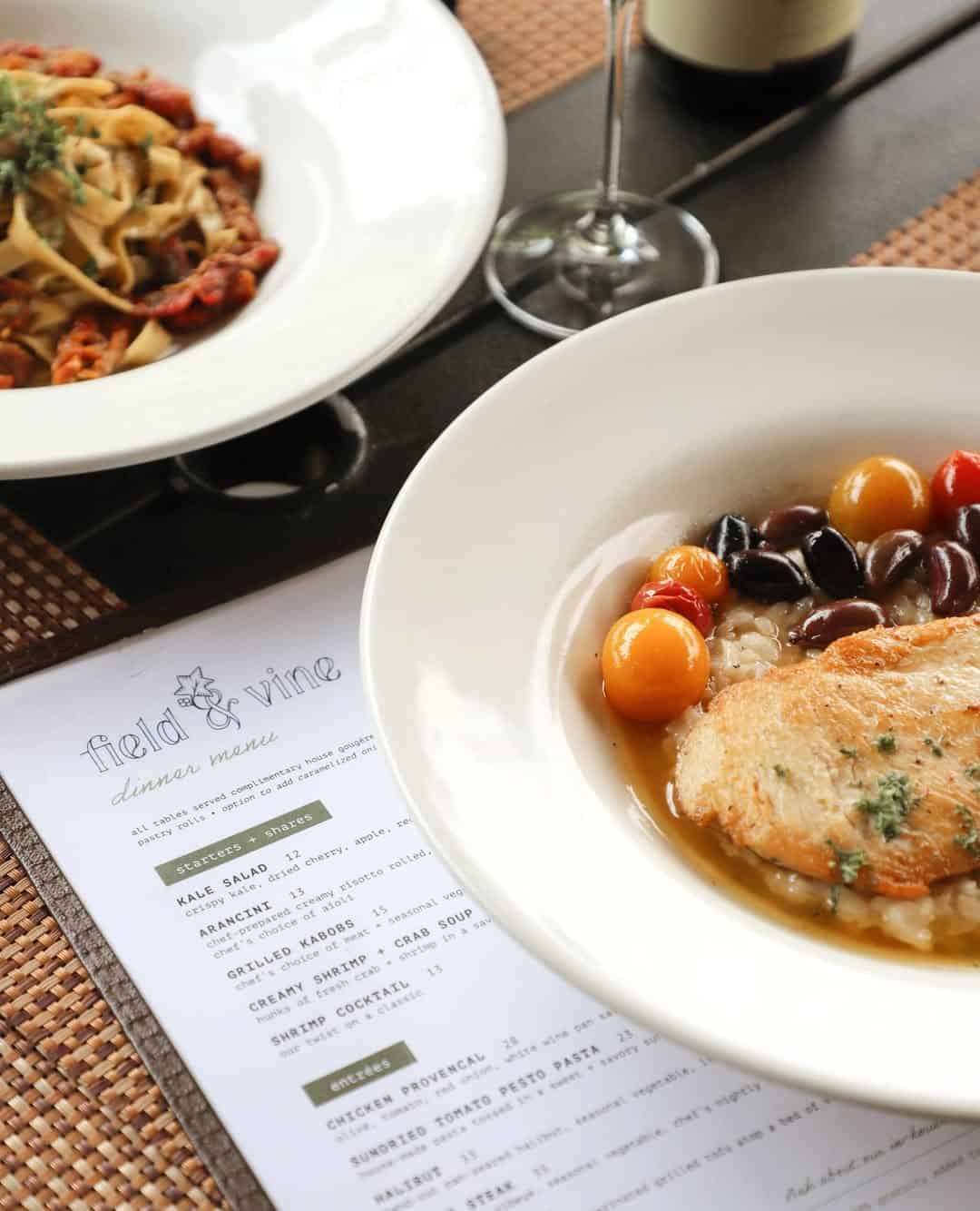 Two plates of pasta and fish with vegetables sit beside a menu, wine glass, and a bottle on a table&mdash;an inviting scene often found at Finger Lakes wineries with restaurants.