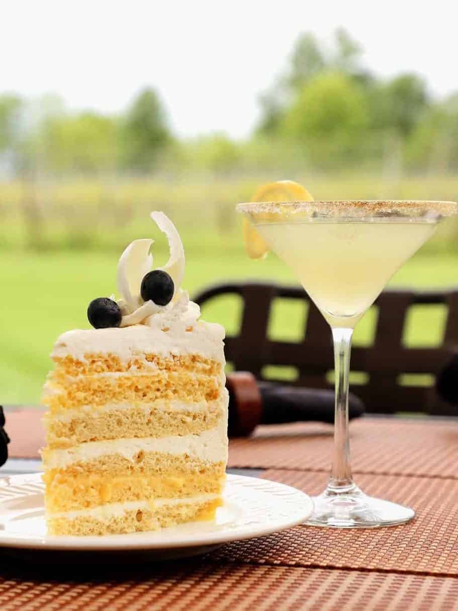 A slice of layered cake with cream and berries sits next to a lemon martini on an outdoor table, evoking the charming atmosphere of Finger Lakes wineries with restaurants.