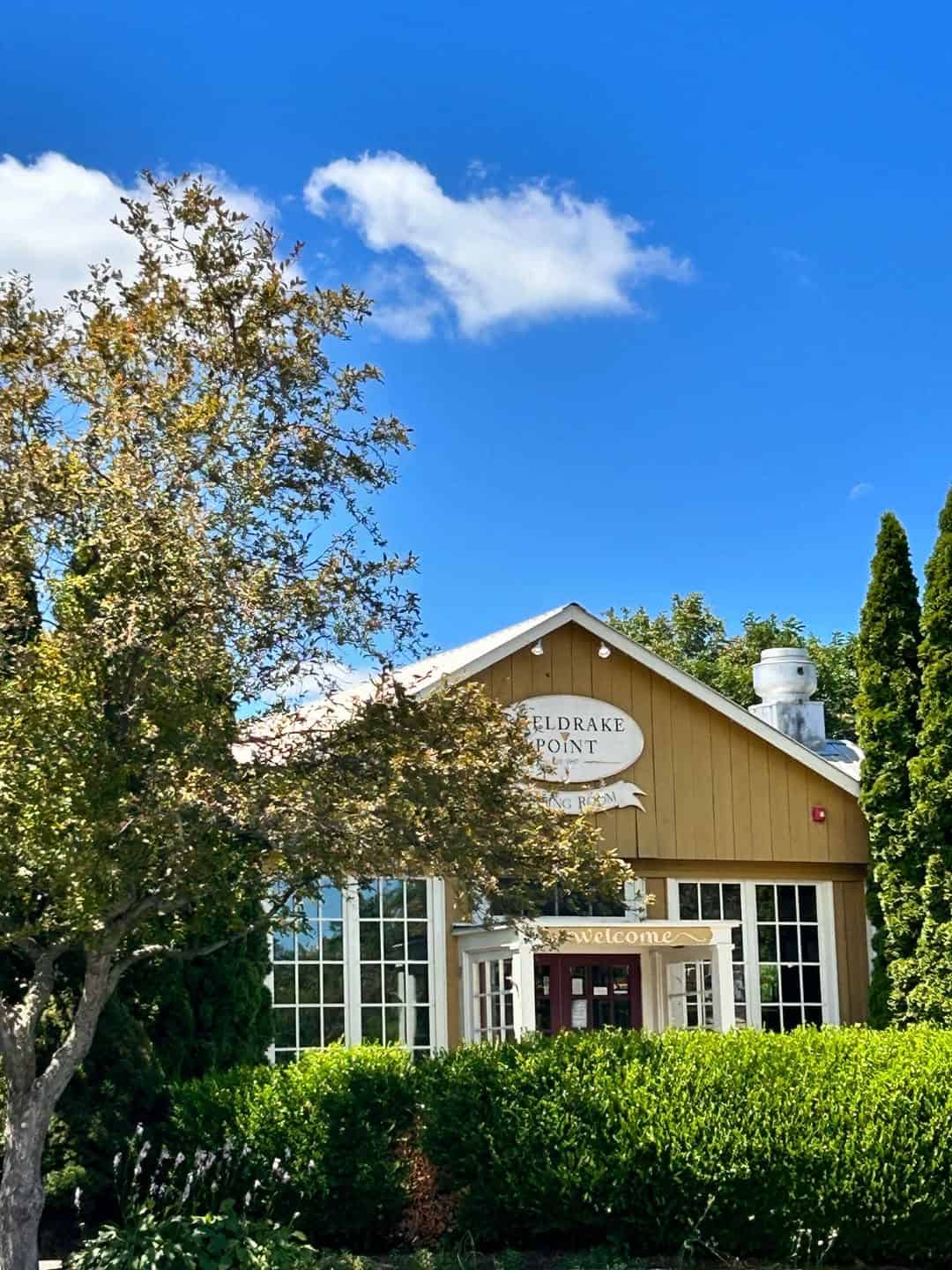 A tan building labeled "Eldridge Point" is surrounded by trees and greenery under a blue sky with clouds, offering the charming ambiance found at many Finger Lakes wineries with restaurants.