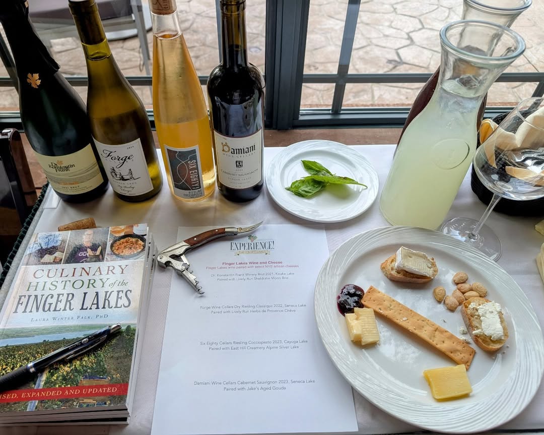 A table with wine bottles from Finger Lakes Wine Tours, cheeses, bread, a book, a menu, and a glass pitcher by a window.