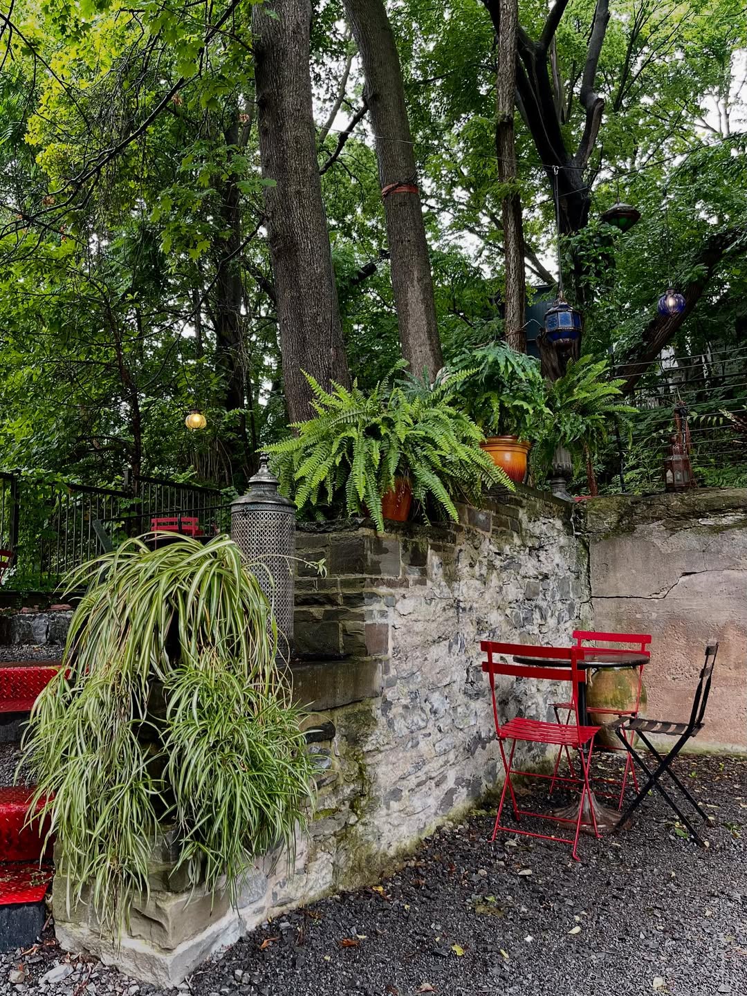 Red bistro table and chairs by a stone wall with potted plants, surrounded by lush green trees—perfect for relaxing after exploring the Best Places to Stay in Finger Lakes Near Wineries.
