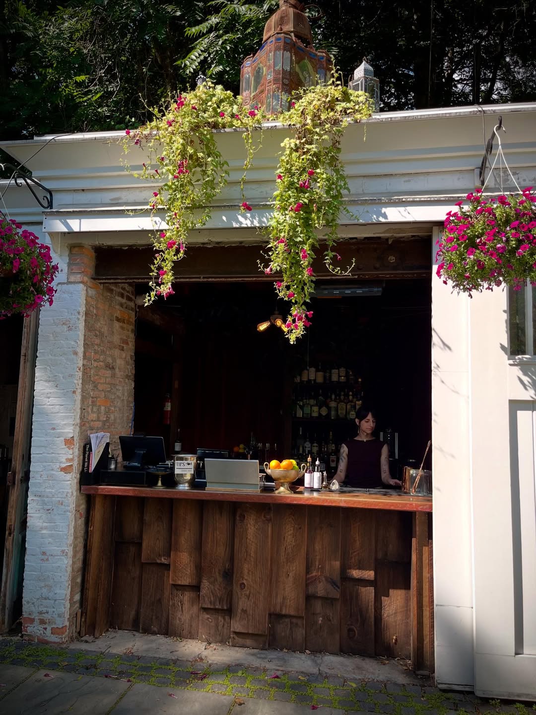 Outdoor bar with a wood counter, hanging flowers, and a bartender standing behind the bar on a sunny day—one of the Best Places to Stay in Finger Lakes Near Wineries for enjoying local charm and scenic relaxation.