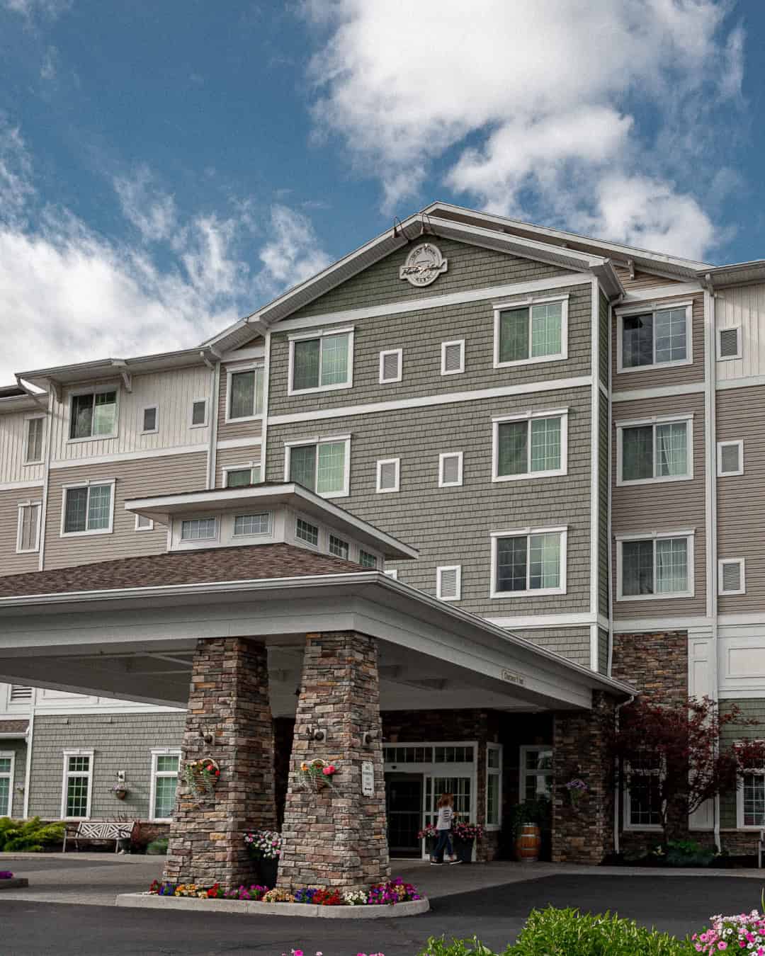 A multi-story hotel with stone pillars, green siding, and flower beds under a cloudy sky—one of the Best Places to Stay in Finger Lakes Near Wineries.