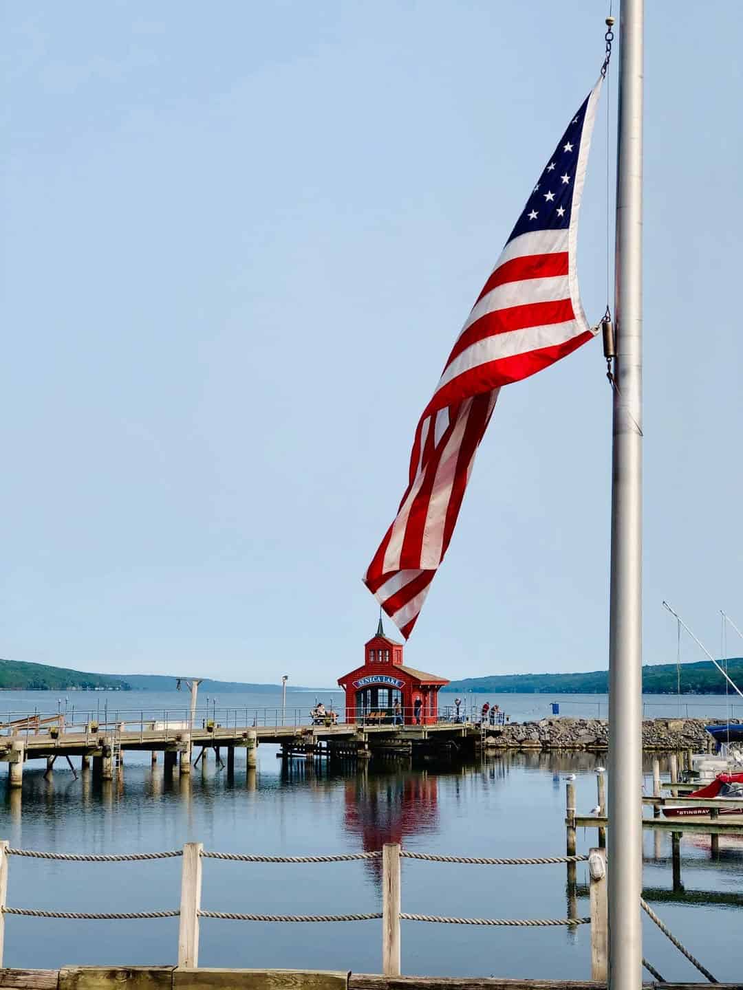 Large American flag waves near a docked red building over calm water with blue sky and distant hills—one of the Best Places to Stay in Finger Lakes Near Wineries for scenic, relaxing getaways.