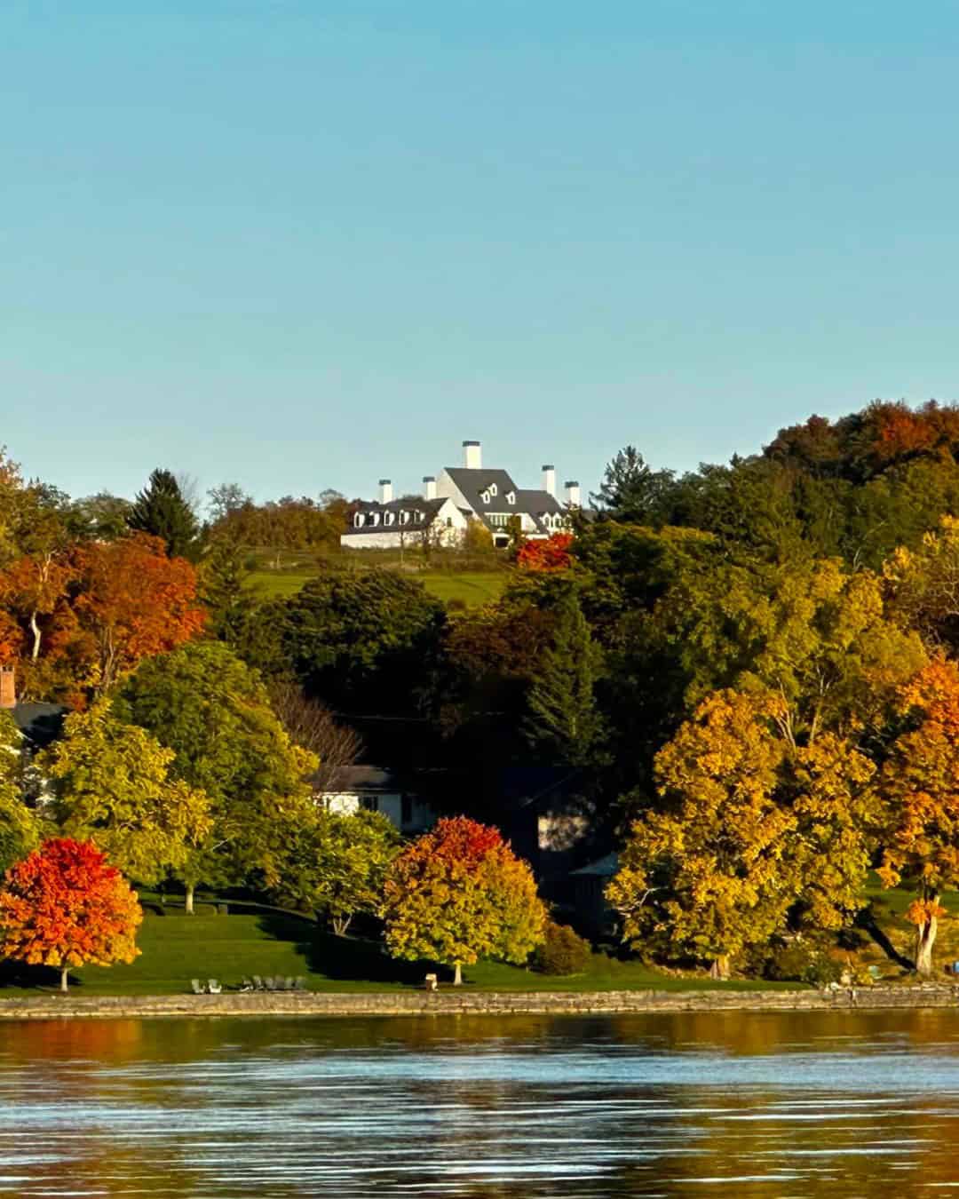 A large house sits atop a hill, surrounded by colorful autumn trees, overlooking a calm body of water—one of the best places to stay in Finger Lakes near wineries.