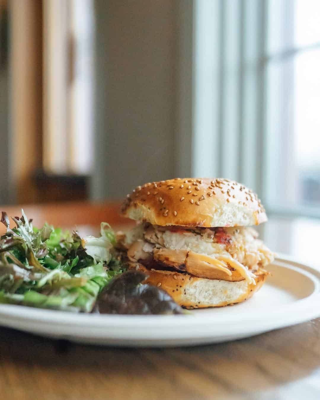 A sesame bun sandwich with turkey and greens, served with a side salad on a white plate by a window&mdash;perfect for enjoying at Finger Lakes wineries with restaurants.