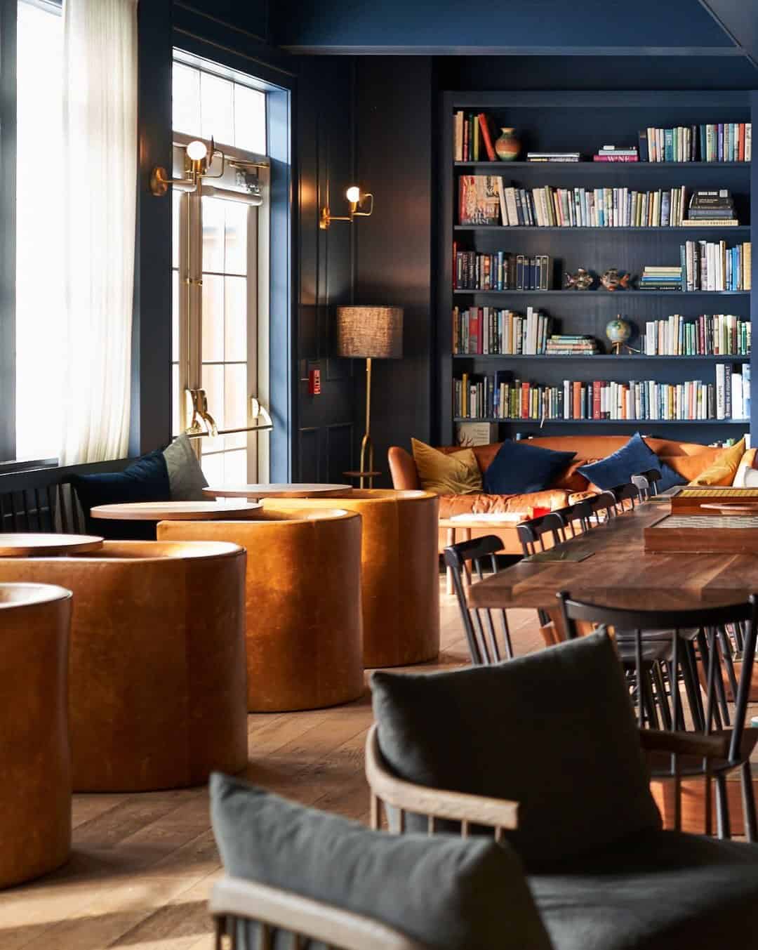 Cozy lounge with wooden tables, cushioned chairs, bookshelves, and large windows with sheer curtains—one of the best places to stay in Finger Lakes near wineries.