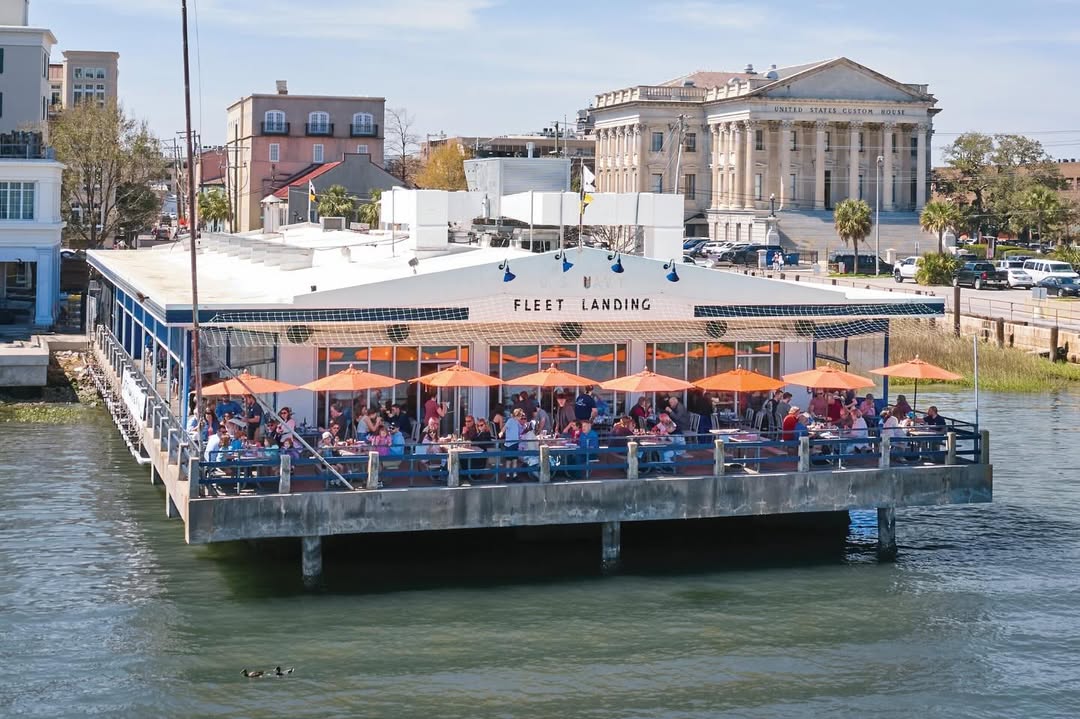 Waterfront restaurant with orange umbrellas and outdoor diners, labeled "Fleet Landing," with city buildings behind—a family-friendly spot that's perfect when searching for things to do in Charleston with kids.