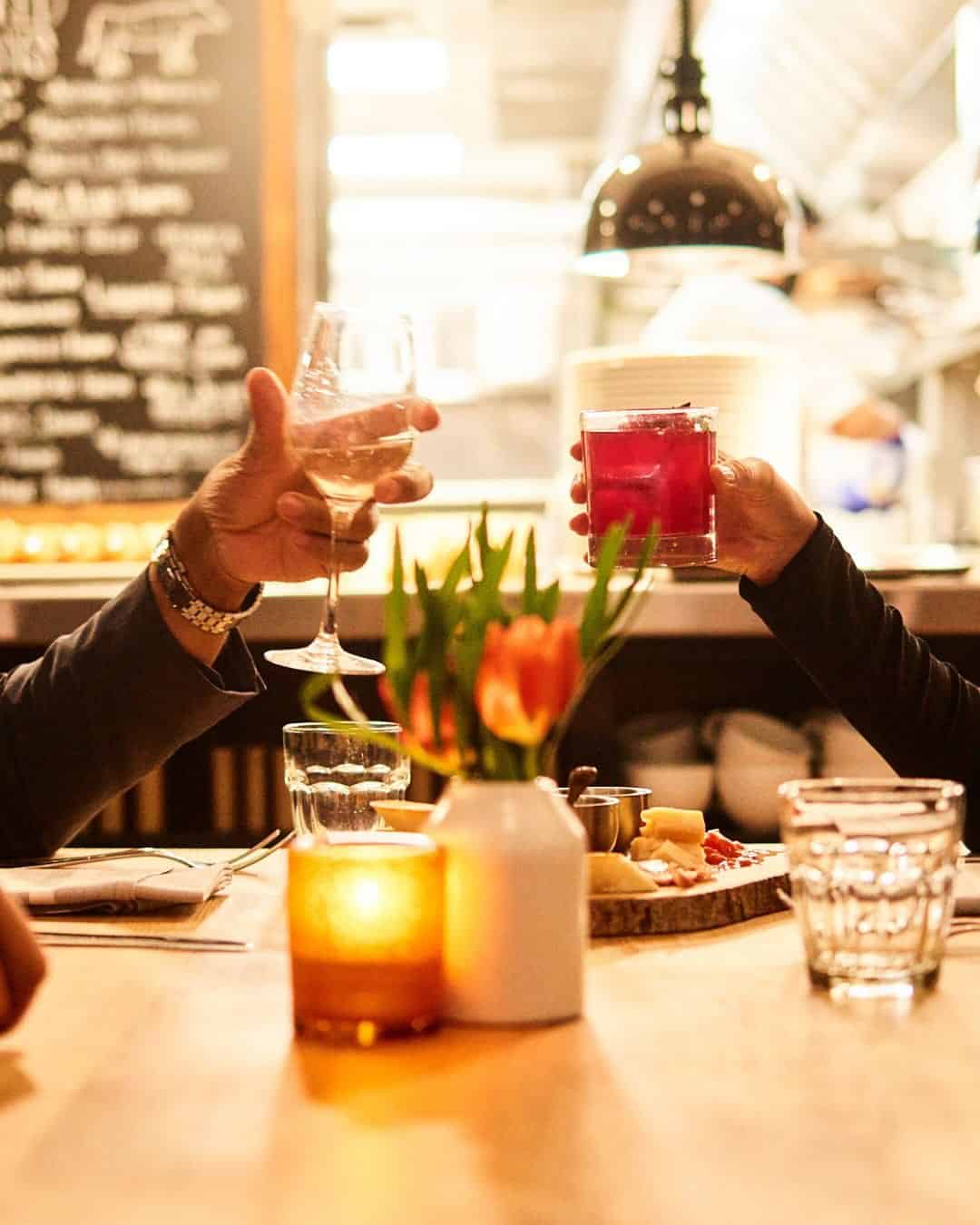 Two people clink drinks over a candlelit restaurant table set with food, flowers, and glasses—an experience to savor after exploring the Best Places to Stay in Finger Lakes Near Wineries.