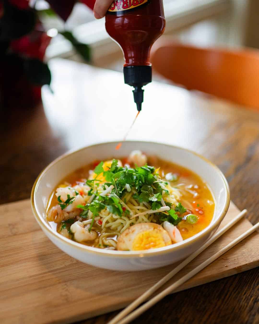 A hand drizzles hot sauce onto a bowl of noodle soup with shrimp, egg, and herbs&mdash;an inviting dish reminiscent of the culinary delights found at Finger Lakes wineries with restaurants. Chopsticks rest beside the bowl.