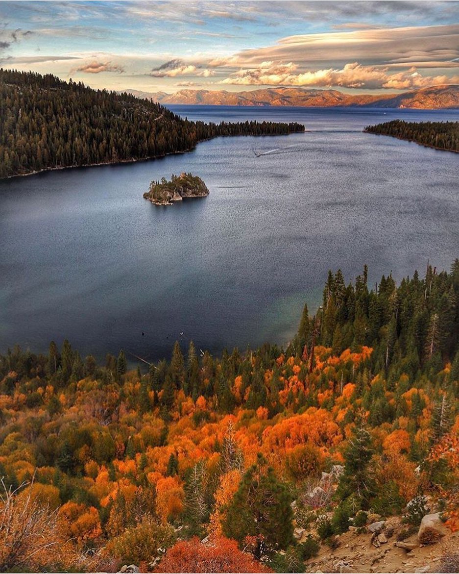 A lake surrounded by forests with autumn foliage, a small island, and distant mountains under a colorful sky—perfect for a family vacation to Lake Tahoe.