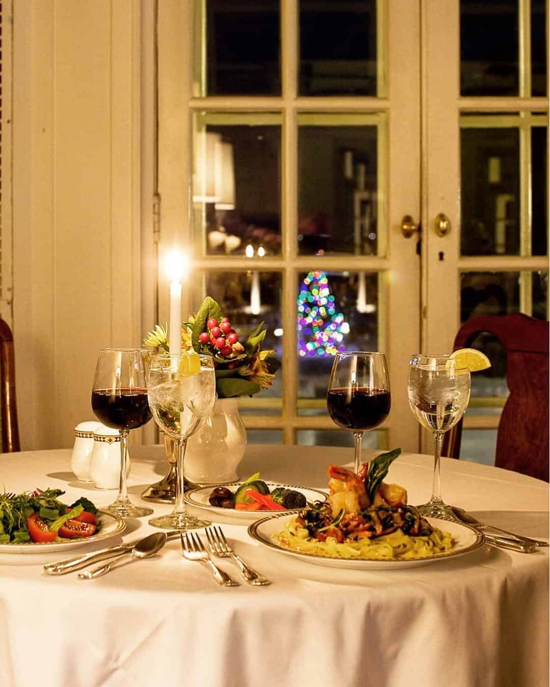 A candlelit dinner table for two with wine, salads, and entrees overlooks a lit Christmas tree outside the window—perfect for those seeking the Best Places to Stay in Finger Lakes Near Wineries.