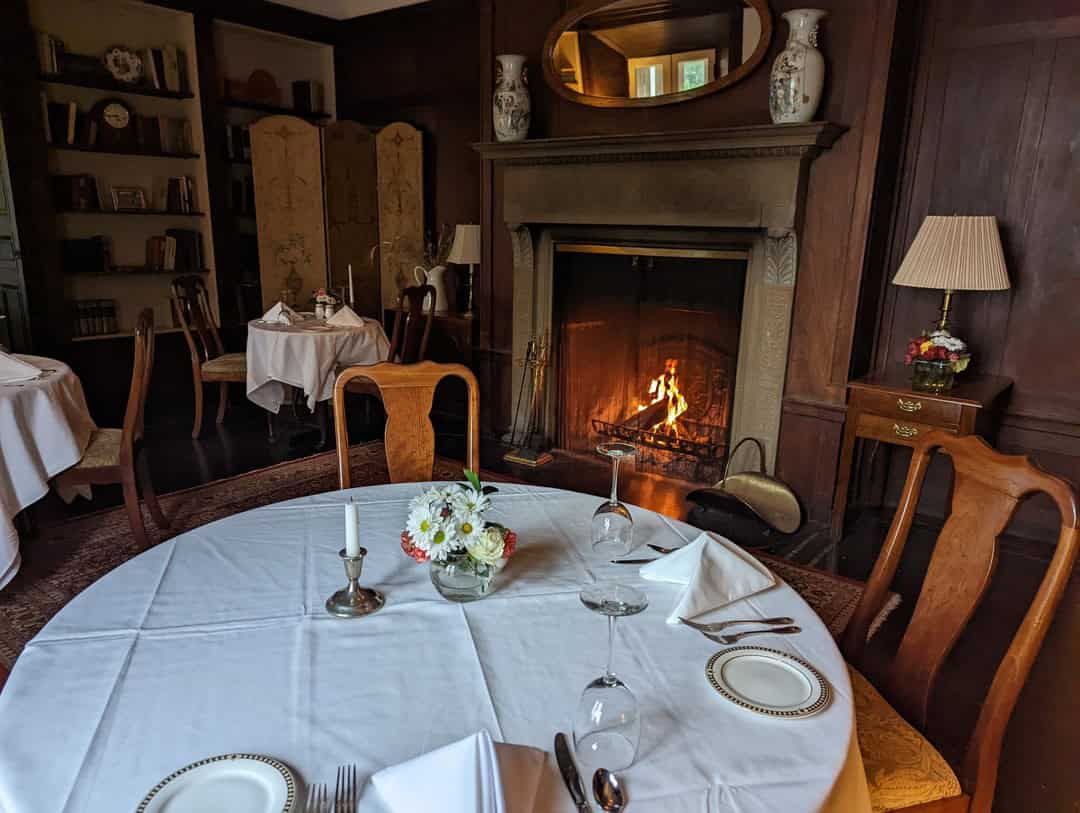 Elegant dining room with a lit fireplace, round table set for four, flowers, and wooden chairs—one of the best places to stay in Finger Lakes near wineries.