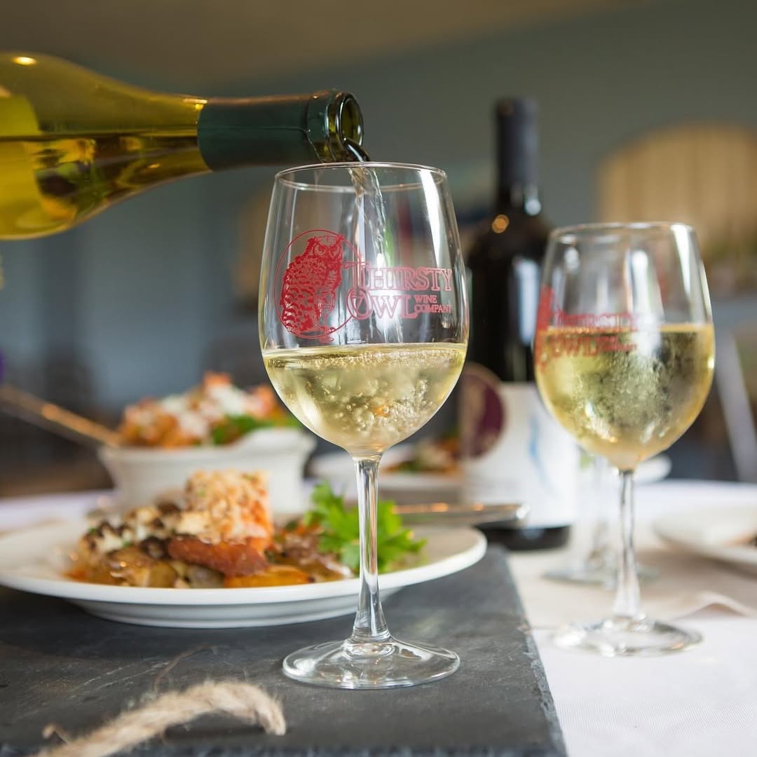 A bottle pours white wine into a glass on a table set with food and another glass of wine, reminiscent of experiences at Finger Lakes wineries with restaurants.