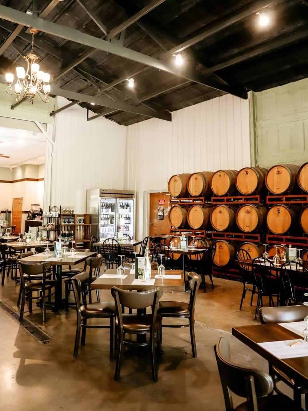 A cozy restaurant with wooden tables and chairs, wine barrels stacked along one wall, and a chandelier above&mdash;reminiscent of the inviting atmosphere found at some Finger Lakes wineries with restaurants.