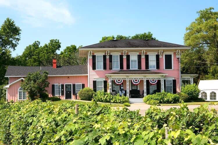 A large pink house with black shutters and patriotic bunting, surrounded by lush greenery and grapevines—one of the best places to stay in Finger Lakes near wineries.