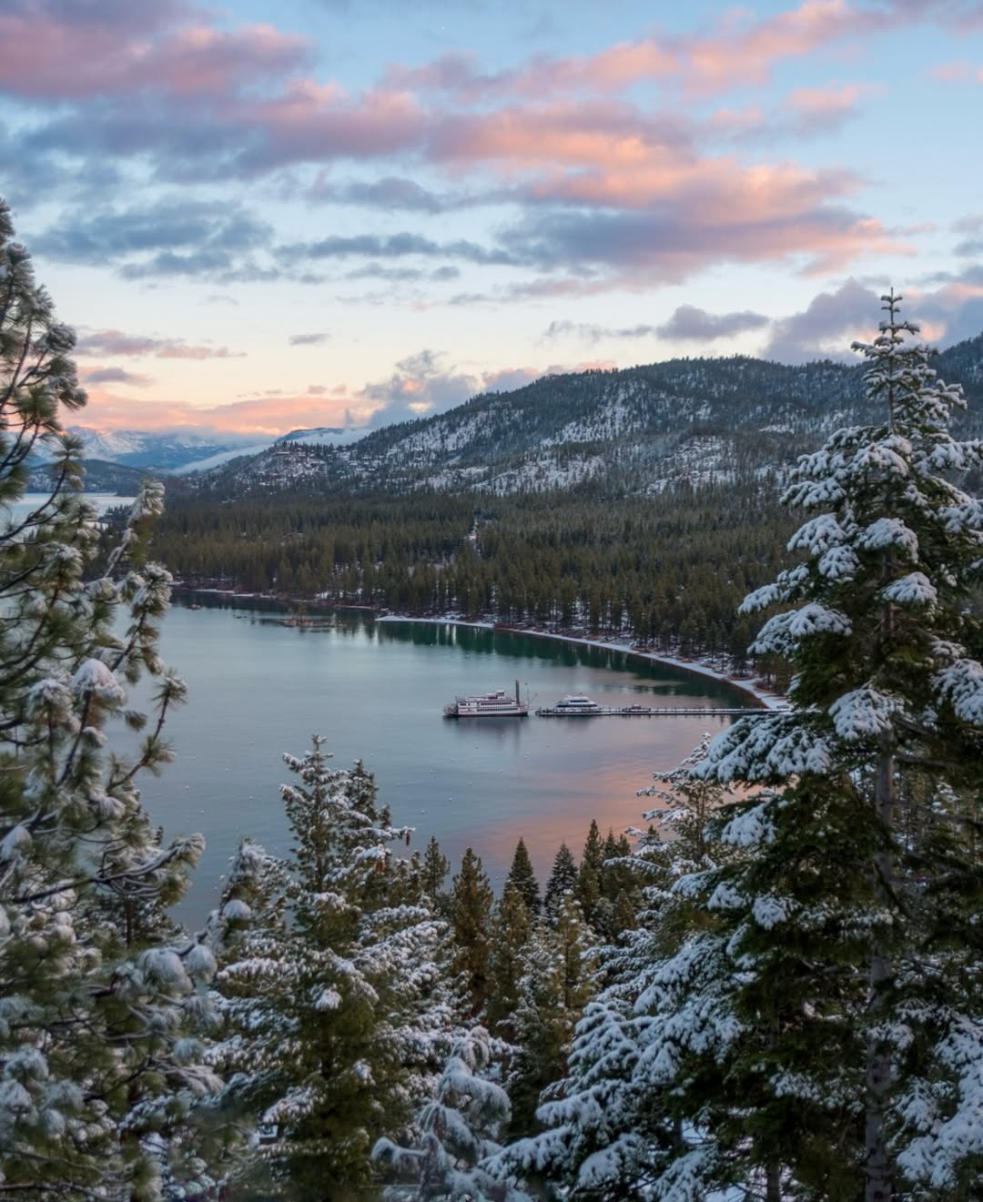 Snow-covered pine trees frame a tranquil lake with boats and mountains in the background at sunset, capturing the serene beauty of a family vacation to Lake Tahoe.