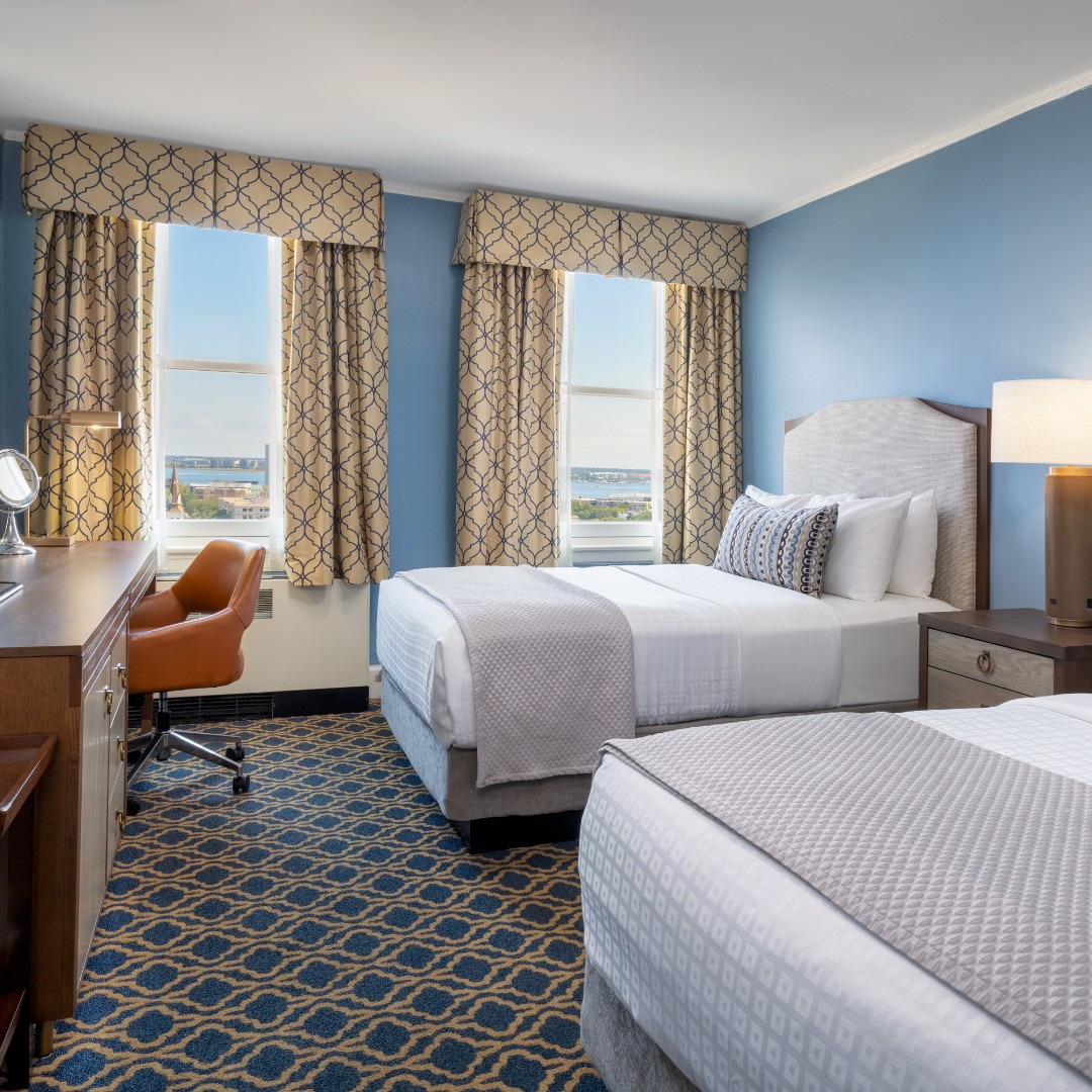 Hotel room with two beds, a desk with an orange chair, and two windows with patterned curtains—perfect for families exploring things to do in Charleston with kids.