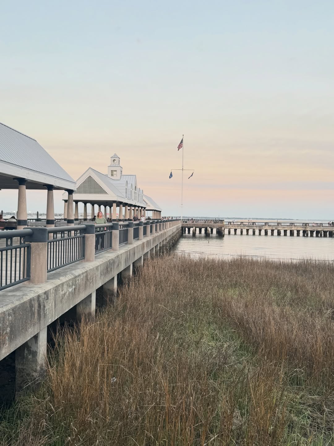 A pier extends over marsh grass toward a pavilion with flags, under a pastel sunset sky by the water—one of the charming things to do in Charleston with kids.