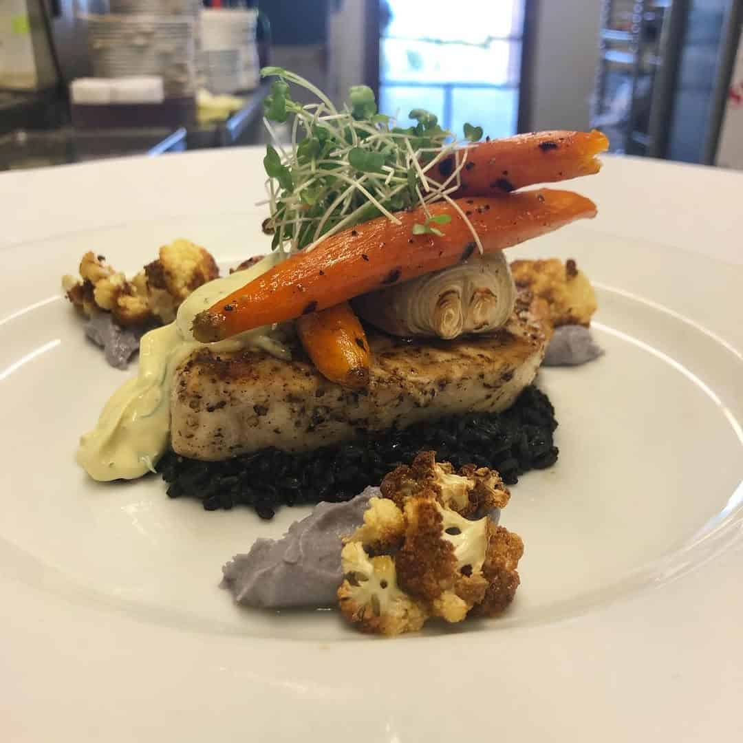 A gourmet dish with grilled fish, black rice, carrots, cauliflower, sauce, and microgreens on a white plate&mdash;perfectly suited for pairing at Finger Lakes wineries with restaurants.