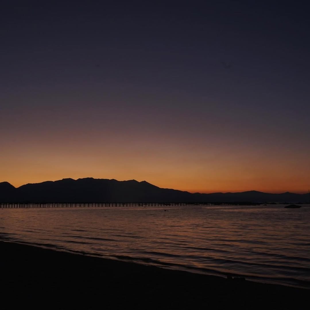 Sunset over calm water with silhouetted mountains and a dark shoreline in the foreground, capturing the peaceful beauty of a family vacation to Lake Tahoe.