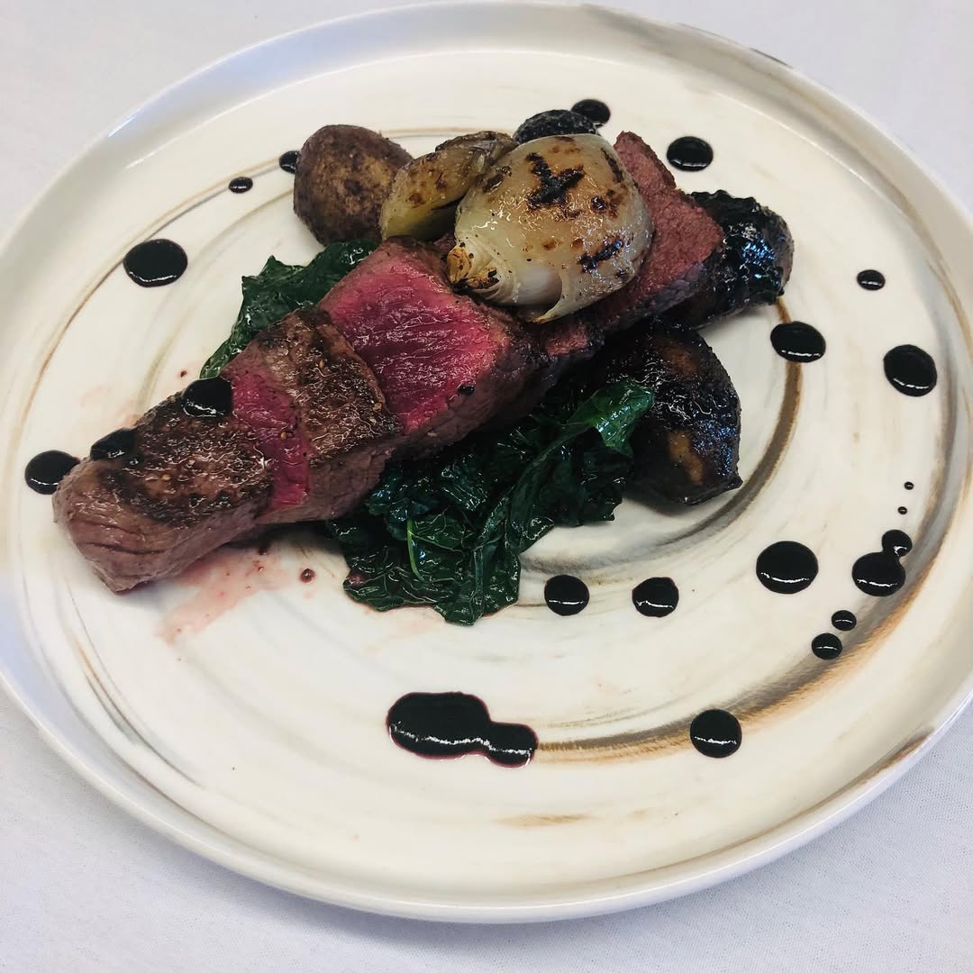 Seared steak on wilted greens with roasted vegetables and balsamic glaze, inspired by dishes found at Finger Lakes wineries with restaurants, elegantly served on a white plate.